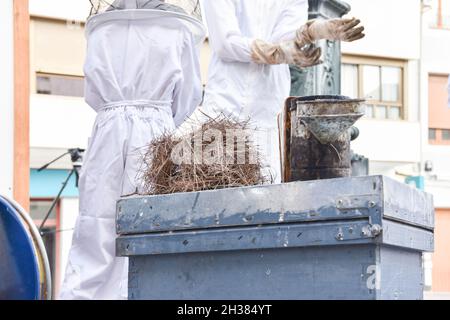 Primo piano di un fumatore di api su un alveare e in background apicoltori che si preparano. Foto Stock