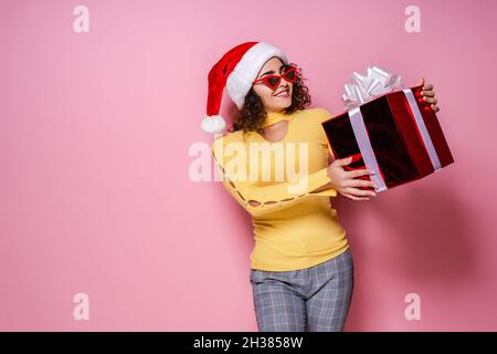 Sorridente ragazza curly nel cappello di Babbo Natale, occhiali da sole tiene giftbox mentre si leva su sfondo rosa Capodanno Foto Stock