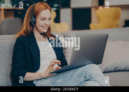 Giovane studentessa positiva di testa di zenzero che ha un webinar online tramite laptop e cuffie in caffetteria Foto Stock