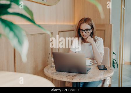 Giovane ragazza bella rossa-capelli freelance donna in auricolari funziona a distanza sul laptop in caffetteria Foto Stock