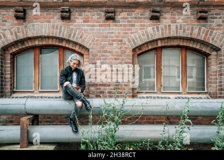 La donna si siede sul tubo vicino a un edificio abbandonato, guarda lo smartphone Foto Stock