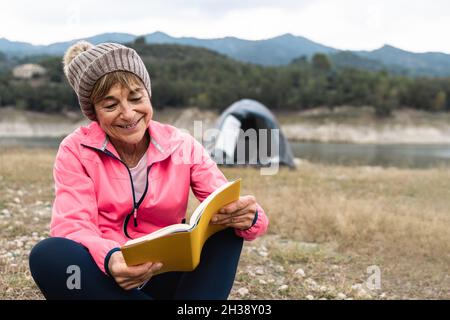 Donna anziana che si diverte a campeggiare con tenda mentre legge un libro all'aperto in un lago - fuoco sul viso Foto Stock