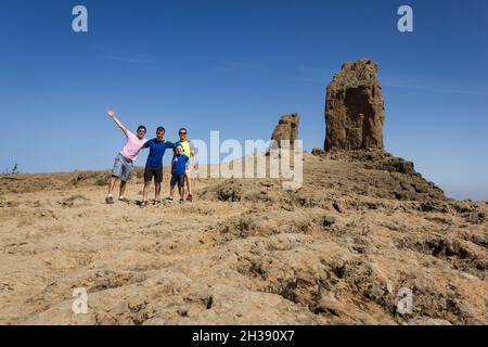 Gruppo di uomini in posa con Roque Nublo sullo sfondo a Gran Canaria. Gli amici si levano in piedi felici dopo l'arrampicata popolare punto di riferimento naturale nelle Isole Canarie Foto Stock