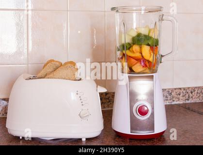 Tostapane bianco con fette di pane e frutta nel frullatore. Kiwi, melone, pezzi di pesca in vaso di vetro. Colazione sana, cucina, elettrodomestici Foto Stock