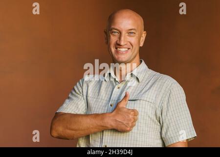 Un uomo di mezza età in una camicia a quadri si alza contro un muro dipinto e guarda la fotocamera con un sorriso amichevole, sorride e mostra un pollice in su Foto Stock