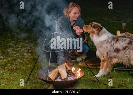 Giovane donna bionda sorridente dà biscotti al suo cane tricolore Australian Shepherd. In inverno, accanto al bollitore per fumare sul fuoco, al buio Foto Stock