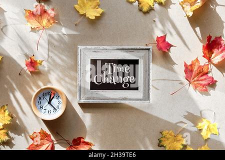 Cambiamento di orario in autunno. Sveglia in legno con un'ora di marcatura con caduta a secco foglie su cartone riciclato. Modifica dell'ora del testo sulla lavagna. Vista dall'alto Foto Stock