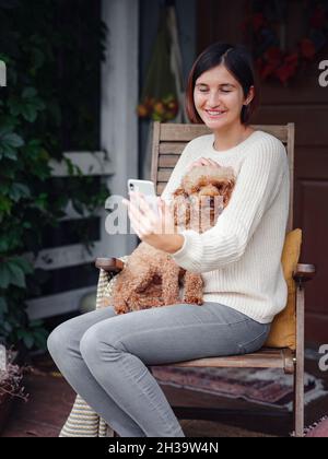 sorridente giovane donna asiatica in maglione beige prende selfie con il suo cucciolo sulla terrazza sul cortile. divertente, accogliente e confortevole stile di vita autunnale Foto Stock