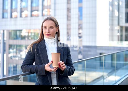 donna d'affari in giacca con caffè in mano si alza sulla terrazza del centro business. profondità di campo poco profonda Foto Stock