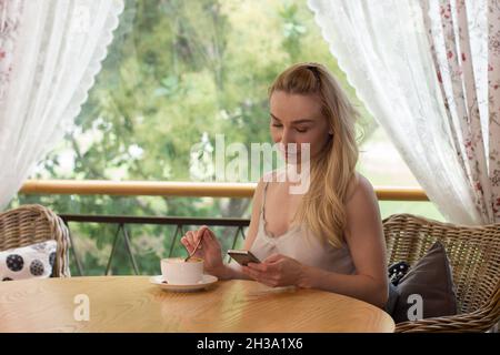 Ragazza che beve caffè. Ragazza guarda il telefono. Balcone per la colazione. Terrazza estiva. Ragazza invia il messaggio di telefono Foto Stock