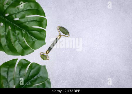 Rullo facciale in pietra di Giada con foglie verdi di monstera. Massaggiatore per il viso. Disposizione piatta. Spazio di copia Foto Stock