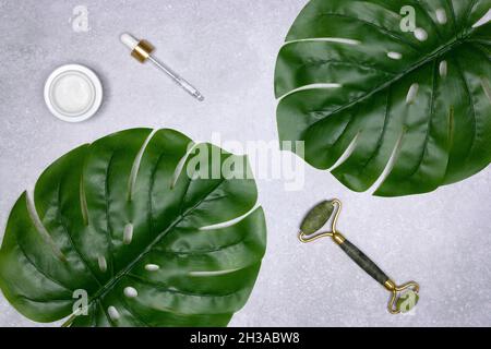 Rullo facciale in pietra di Giada, vasetto di crema, contagocce con foglie verdi di monstera. Massaggio viso e prodotti per la cura della pelle. Disposizione piatta Foto Stock