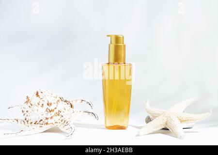 Bottiglia trasparente di olio giallo-arancione per proteggere il sole, l'abbronzatura o i capelli. Fondo bianco con olio cosmetico e conchiglie. Cosmetici per la cura estiva. Foto Stock