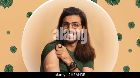 Sorridente ragazzo arabo millenario che mostra spalla con aiuto banda dopo la vaccinazione su sfondo beige Foto Stock