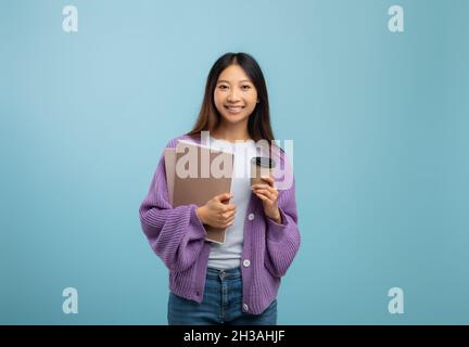 Eccitata studentessa asiatica che tiene taccuini e caffè per andare, avendo pausa durante lo studio, sfondo blu Foto Stock
