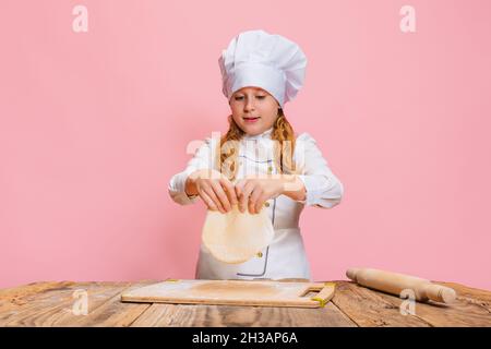 Ritratto di piccola bella ragazza in uniforme cuoco bianco ed enorme cappello dello chef a cucina dei bambini isolato su sfondo rosa studio. Foto Stock