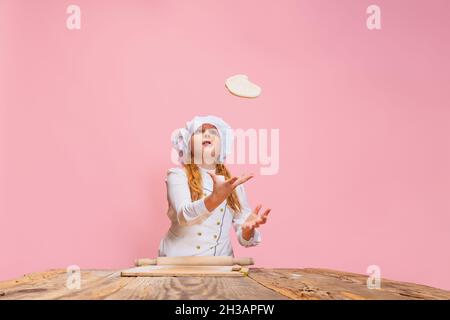 Ritratto di piccola bella ragazza in uniforme cuoco bianco ed enorme cappello dello chef a cucina dei bambini isolato su sfondo rosa studio. Foto Stock