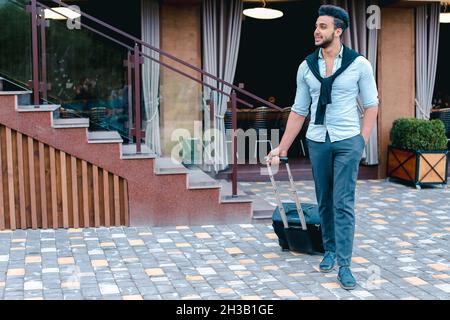 Il giovane uomo d'affari arabo Guy studente è venuto al ristorante con Suitcase in mano, sorridendo a Camera, si ferma e guarda via. Foto Stock