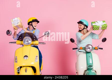 Foto di eccitati marito anziano moglie guida moto vestiti caschi tenendo presenti scatole isolato rosa sfondo di colore Foto Stock