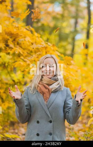 Una donna sorridente gettò un mazzo di foglie in un parco contro uno sfondo di alberi. Autentica donna di 40 anni senza ritocchi Foto Stock