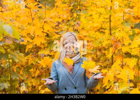 Una donna sorridente gettò un mazzo di foglie in un parco contro uno sfondo di alberi. Autentica donna di 40 anni senza ritocchi Foto Stock