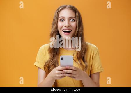 Girata in vita di giovane ragazza allegra ed eccitata con acconciatura ondulata sorridente ampiamente stupito e stupito reagire alla presa impressionante dello smartphone Foto Stock