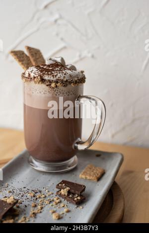 S'Mores cioccolata calda con cracker graham, panna montata e polvere di cacao in tazza trasparente su piastra rettangolare in ceramica grigia macchiata fatta a mano. Foto Stock