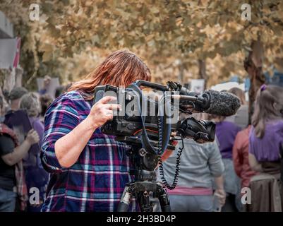 Videografo televisivo femminile con macchina fotografica professionale che filma a protesta affollata il giorno d'autunno - primo piano Foto Stock