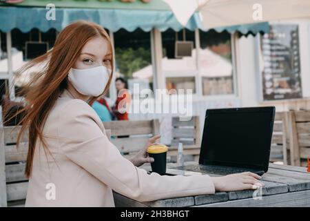 Donna d'affari Redhead che indossa la maschera per la protezione dai virus lavorando sul laptop all'aperto. Giovane Businesswoman che lavora su un notebook all'esterno. Foto Stock