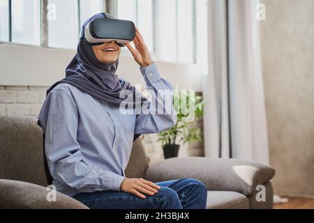 Gioiosa donna araba seduta su un divano, con un casco VR e sorridente Foto Stock
