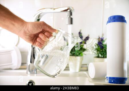 Uomo che riempie una caraffa di vetro da un rubinetto di acqua purificata con un sistema di osmosi del lavello della cucina domestica con filtri intorno. Vista frontale. Compo. Orizzontale Foto Stock