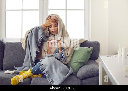 Giovane donna malata, avvolta in coperte, che guarda il termometro dopo aver preso la sua temperatura Foto Stock