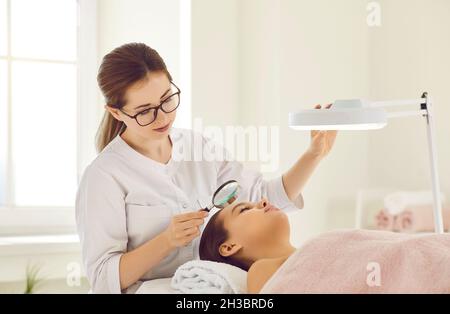 Donna beautician tenendo piccola lente d'ingrandimento guardando attraverso di esso esaminare la condizione della pelle. Foto Stock