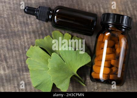 Ginkgo biloba .vaso di vetro marrone con pillole omeopatiche con estratto di ginkgo e foglie di ginkgo su sfondo di legno . Medicina alternativa e omeopatia Foto Stock