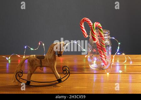 Decorazione di Natale - piccolo giocattolo di legno a cavallo e caramello caramelle di Capodanno in un vaso di vetro intrugiato con una luminosa ghirlanda festiva su un tavolo Foto Stock