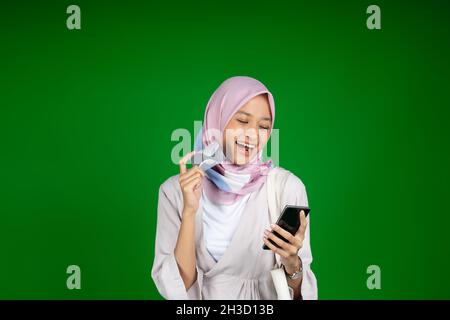 Donna eccitata in hijab usando il telefono cellulare mentre tiene la carta atm Foto Stock