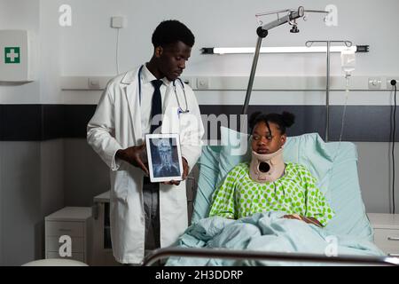 Paziente afroamericano che indossa collo cervicale poggiato a letto recupero dopo chirurgia medica. Medico terapista che tiene una tavoletta con radiografia ossea sullo schermo che lavora in ospedale reparto Foto Stock