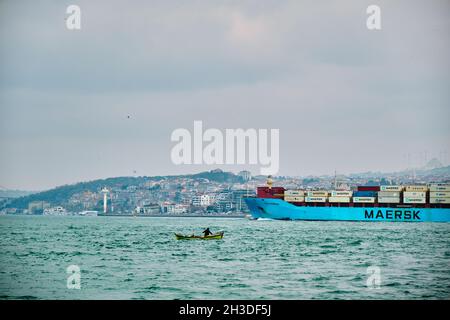 Enorme nave di trasporto (MAERSK) che passa attraverso a istanbul bosforo durante la giornata colta e piovosa con piccola barca da pesca e sfondo ponte Foto Stock