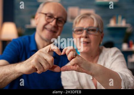 Coppia anziana che crea un segno di forma cardiaca con le mani a casa. Sposato persone anziane in amore facendo romantico simbolo mentre si guarda la macchina fotografica in soggiorno. Marito e moglie mostrano figura di affetto Foto Stock