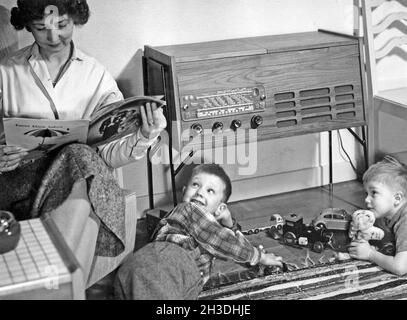 Negli anni '50. Una madre è vista seduta leggendo una rivista nella sua casa, mentre i suoi figli stanno giocando con le automobili giocattolo sul pavimento. Si vede un tipico radio-recordplayer degli anni '50 con un armadietto in legno. Svezia 1957 Foto Stock