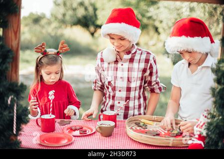 Bambini felici che si preparano per Natale. Due ragazzi e bambina in biscotti di pan di zenzero di colore santa Hat, bevendo cioccolata calda fuori divertirsi. Deco bambini Foto Stock