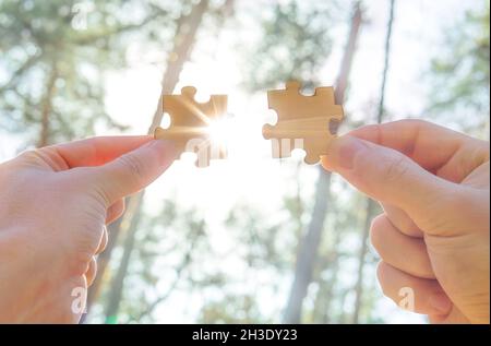 Puzzle contro uno sfondo di foresta. Due mani che collegano due pezzi di puzzle con la luce del sole e raggi che passano attraverso. Lavoro di squadra e coopera d'affari Foto Stock