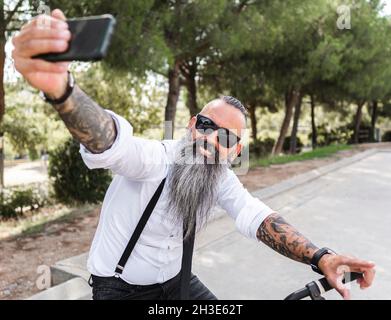 Positivo uomo con bearded in occhiali da sole e camicia bianca che prende autoritratto su smartphone mentre si siede in bicicletta nel parco con alberi Foto Stock