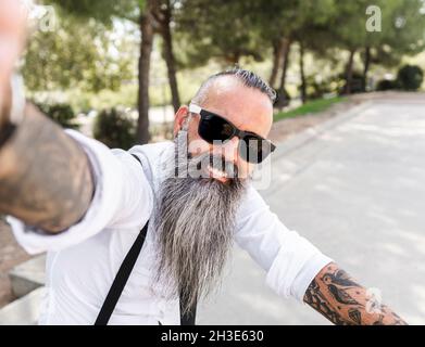 Positivo uomo con bearded in occhiali da sole e camicia bianca che prende autoritratto su smartphone mentre si siede in bicicletta nel parco con alberi Foto Stock