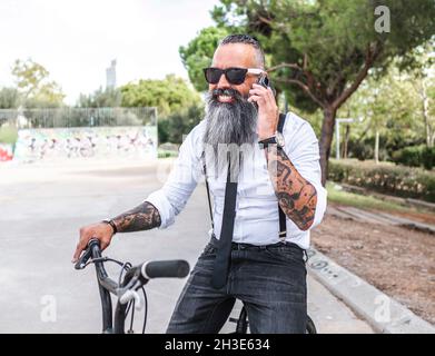 Uomo con occhiali da sole e camicia bianca che parla sullo smartphone mentre si siede in bicicletta nel parco con alberi Foto Stock