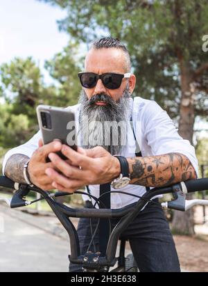 Uomo con occhiali da sole e camicia bianca che naviga sullo smartphone mentre si siede in bicicletta nel parco con alberi Foto Stock