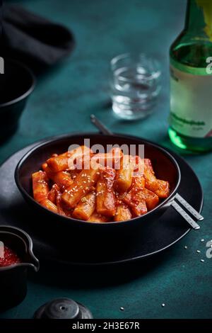 Mucchio di torta di riso speziato tteokbokki fritto con semi di sesamo servito in una ciotola nera sul tavolo con bevanda alcolica Foto Stock
