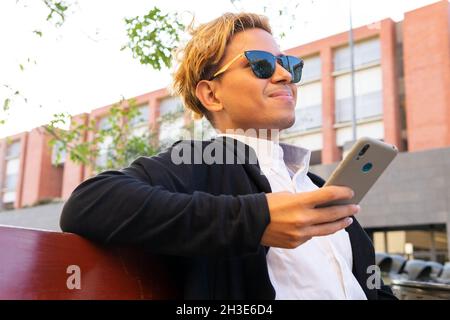 Allegro giovane maschio in occhiali da sole e elegante vestito di messaggi di testo sul cellulare mentre si siede sulla strada vicino edificio in città Foto Stock