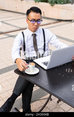 Giovane imprenditore asiatico maschio wistful con tazza di bevanda calda e netbook guardando lo schermo nel tavolo urbano della caffetteria alla luce del giorno Foto Stock