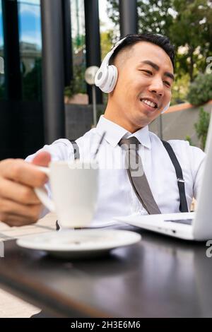 Contenuto giovane imprenditore asiatico maschile in cuffia wireless con tazza di caffè durante la video chat sul laptop in strada caffetteria Foto Stock